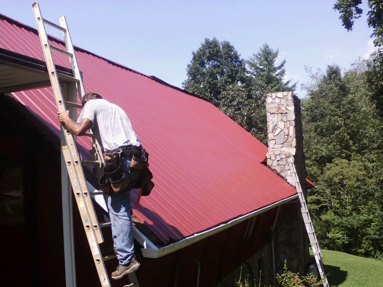 Roofer on a ladder beside a red metal roof and stone chimney — Soffit & Fascia Repair by Roofing Brothers in Oklee